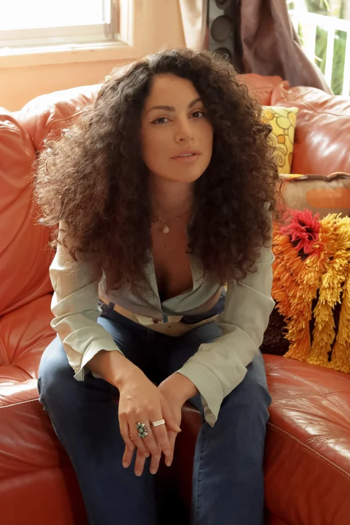 Dark curly-haired sitting on couch near a bookshelf of records.