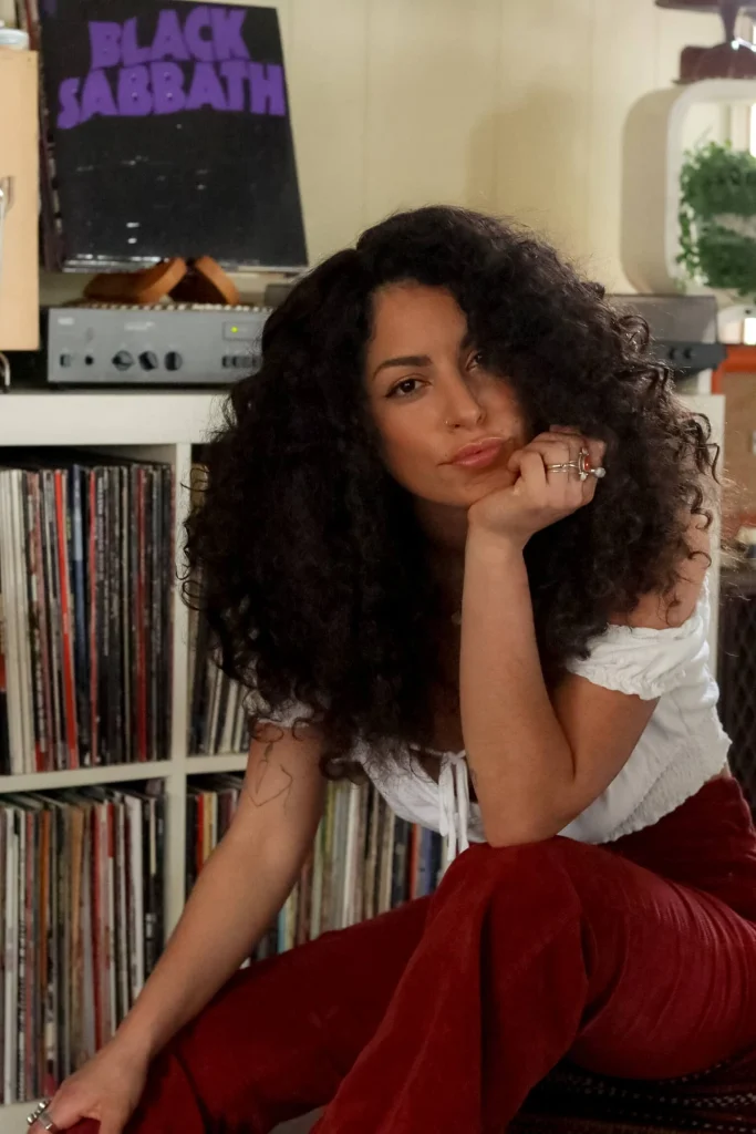 Dark curly-haired sitting on couch near a bookshelf of records.