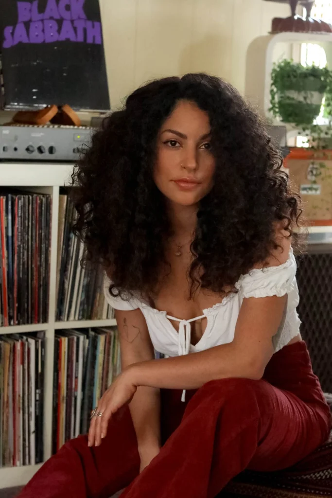Dark curly-haired sitting on couch near a bookshelf of records.