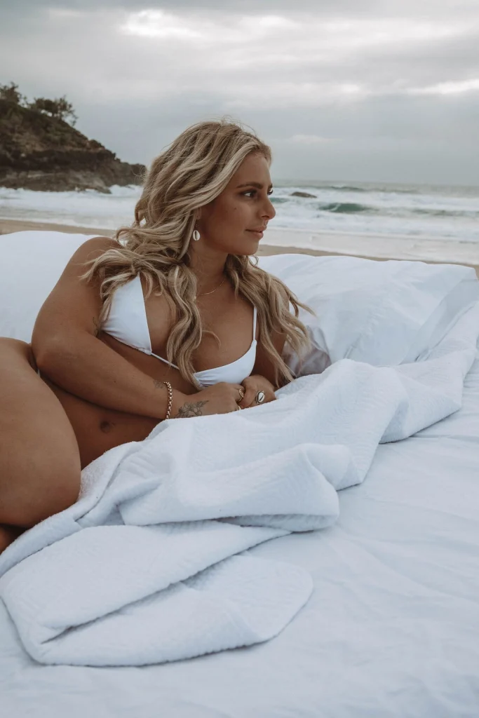 Blond woman wearing a bikini laying on bedding on the beach.