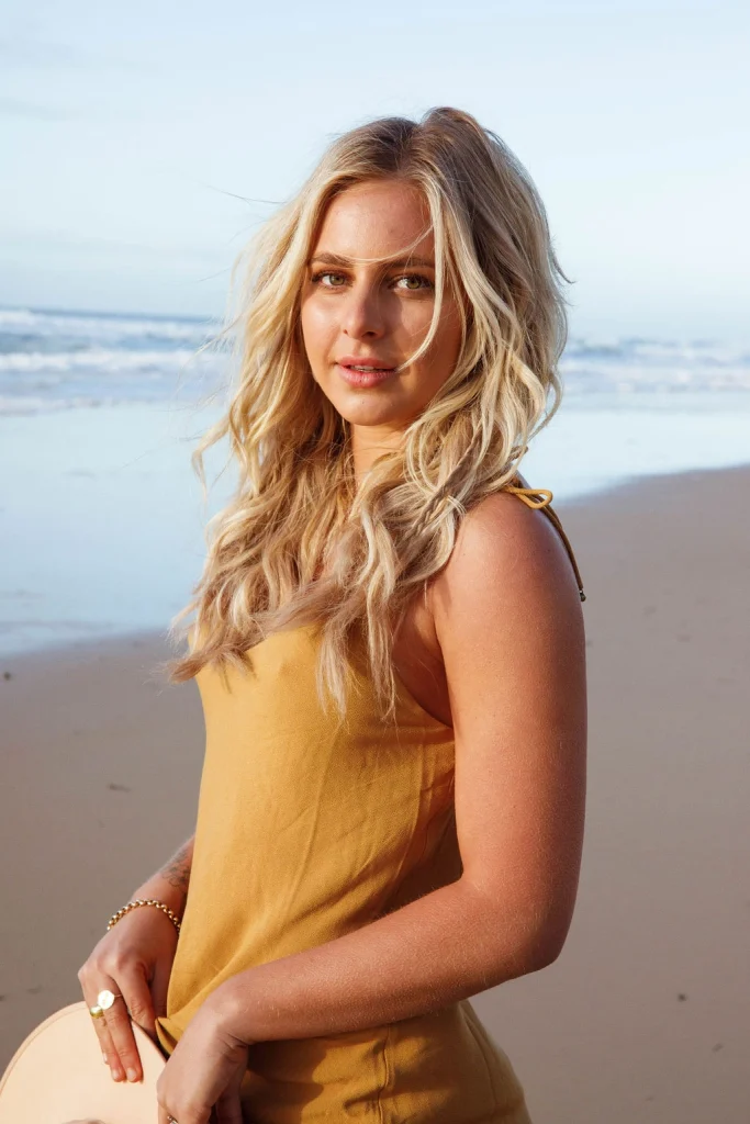 Blond woman wearing a mustard colored dress and jewellery on the beach.