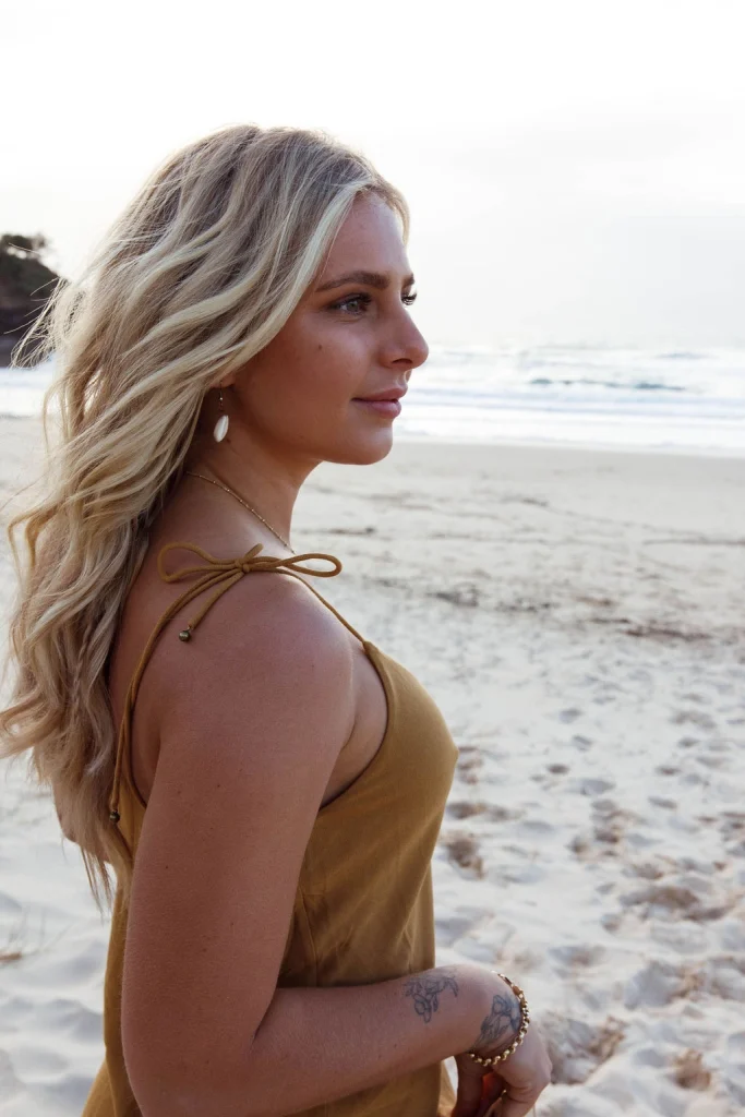 Blond woman wearing a mustard colored dress and jewellery on the beach.