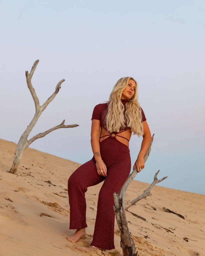 Blond woman in a red jumpsuit holding a tree on sand dunes.