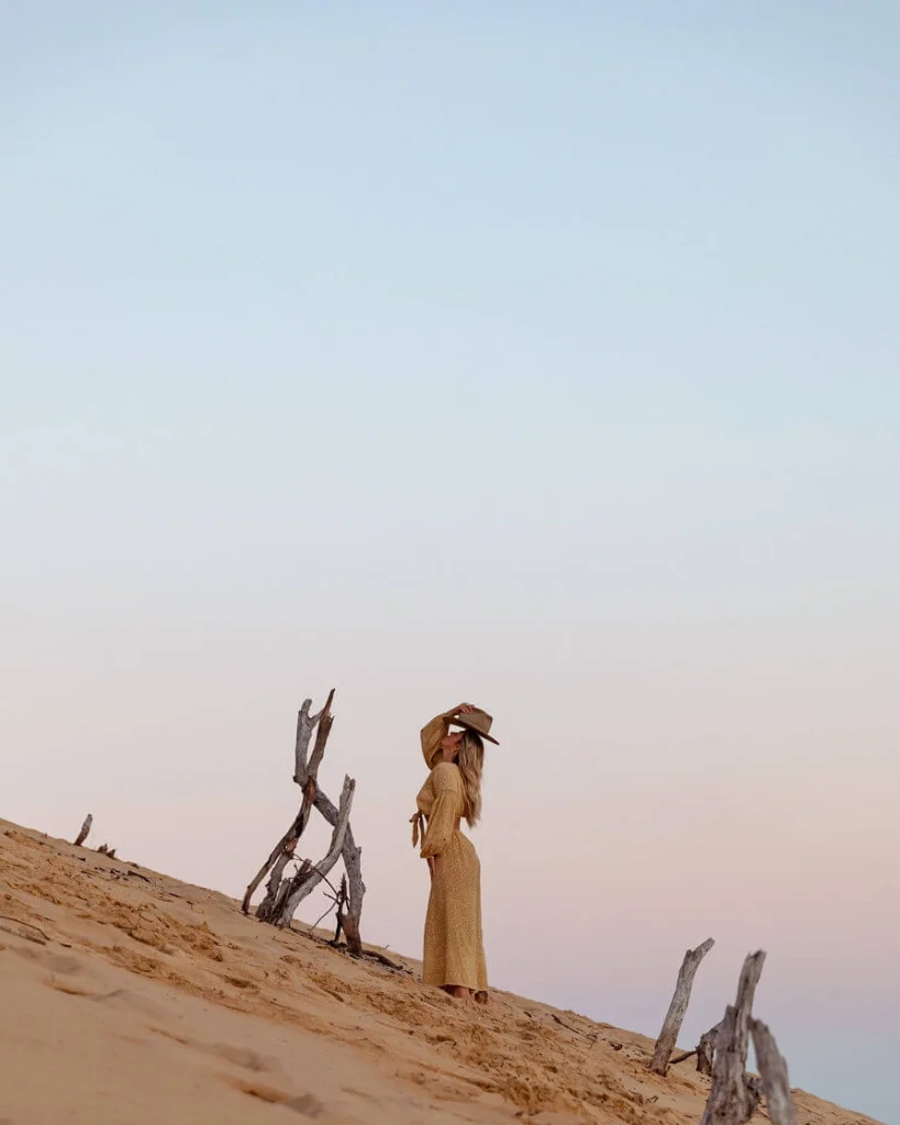 Blond woman in a hat and mustard colored dress holding a tree on sand dunes.