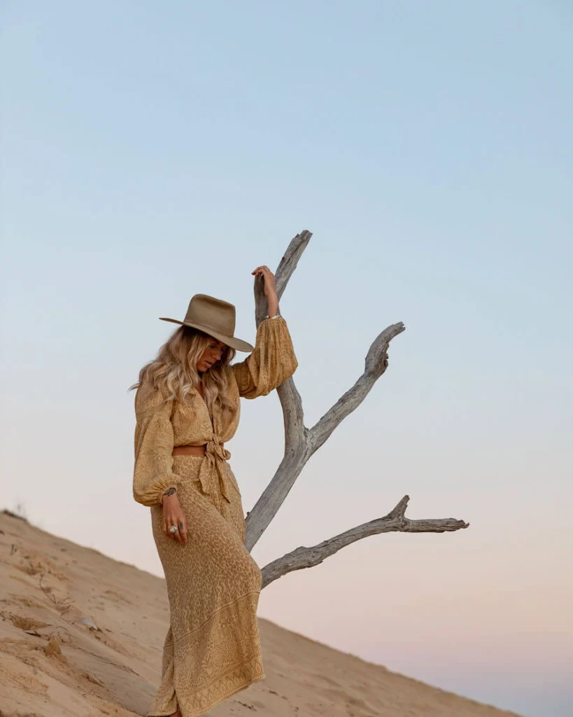Blond woman in a hat and mustard colored dress holding a tree.