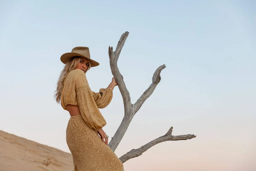Blond woman in a hat and mustard colored dress holding a tree.
