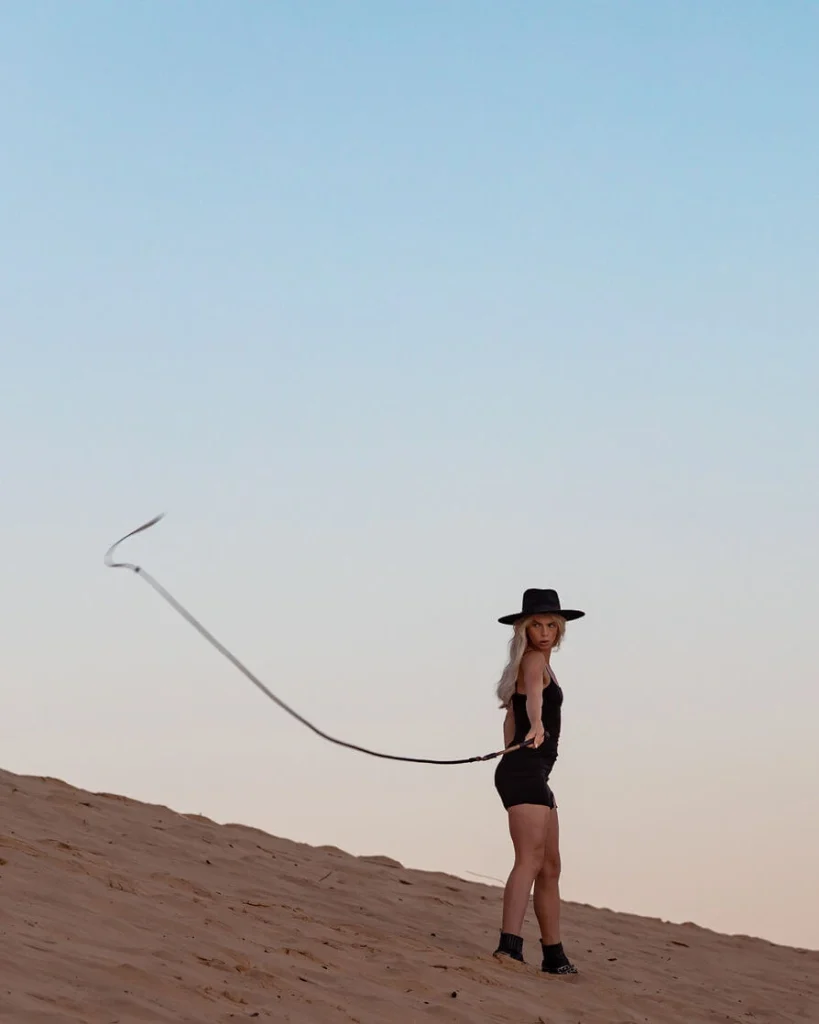 Blond woman in a black hat on sand dunes holding a whip.