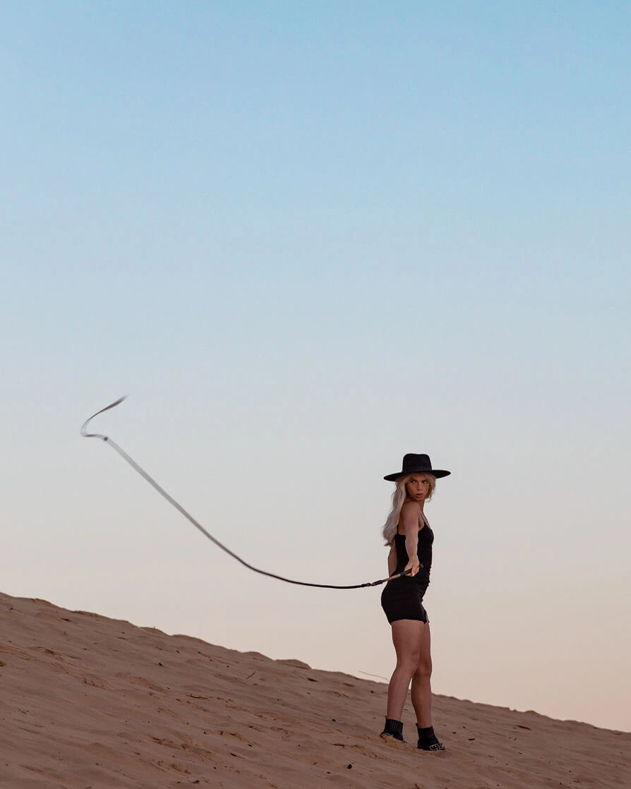 Blond woman in a black hat on sand dunes holding a whip.