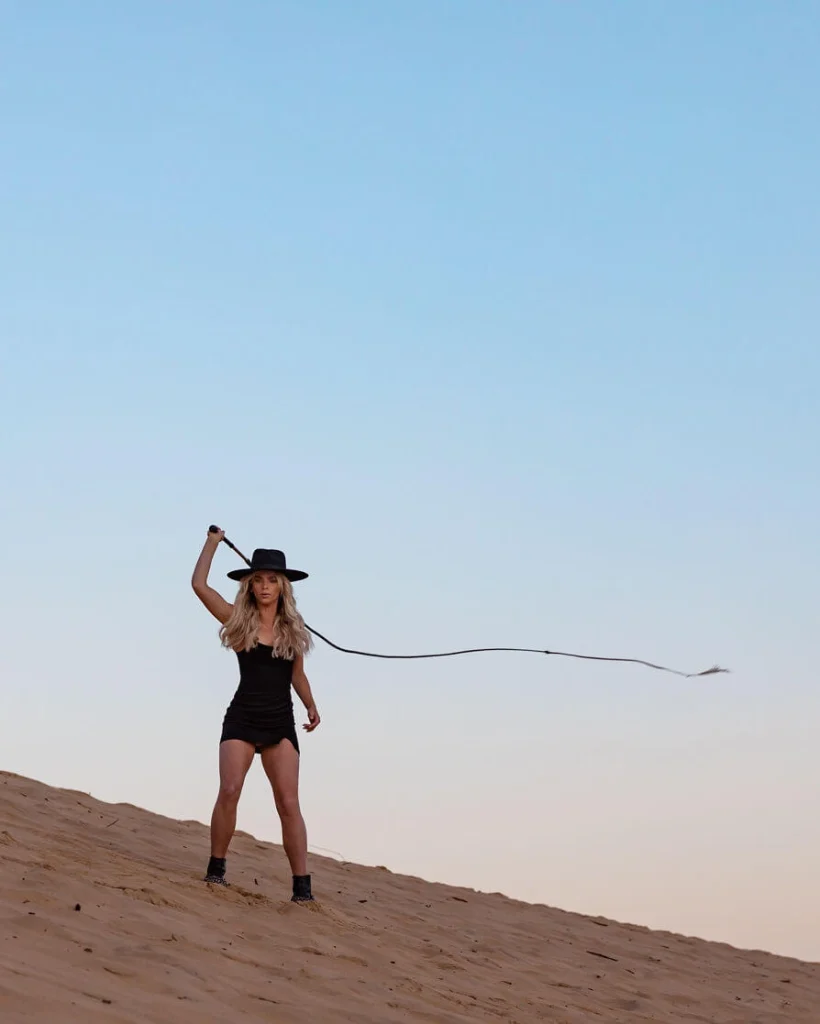 Blond woman in a black hat on sand dunes holding a whip.