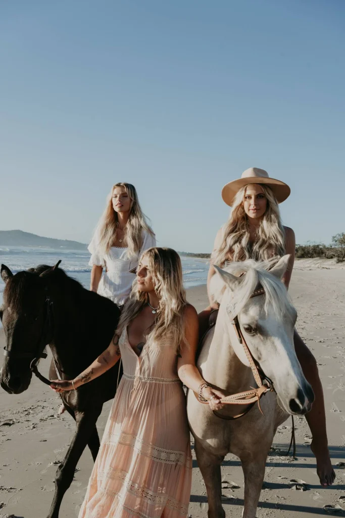 Two blond women on horses on the beach, and another walking in the centre.