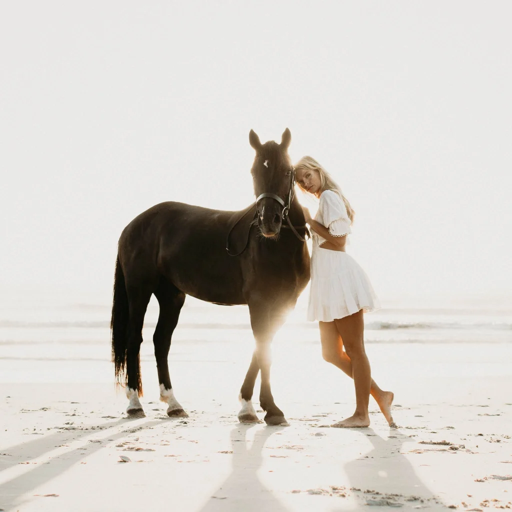 Blond woman standing next to dark horse on the beach.