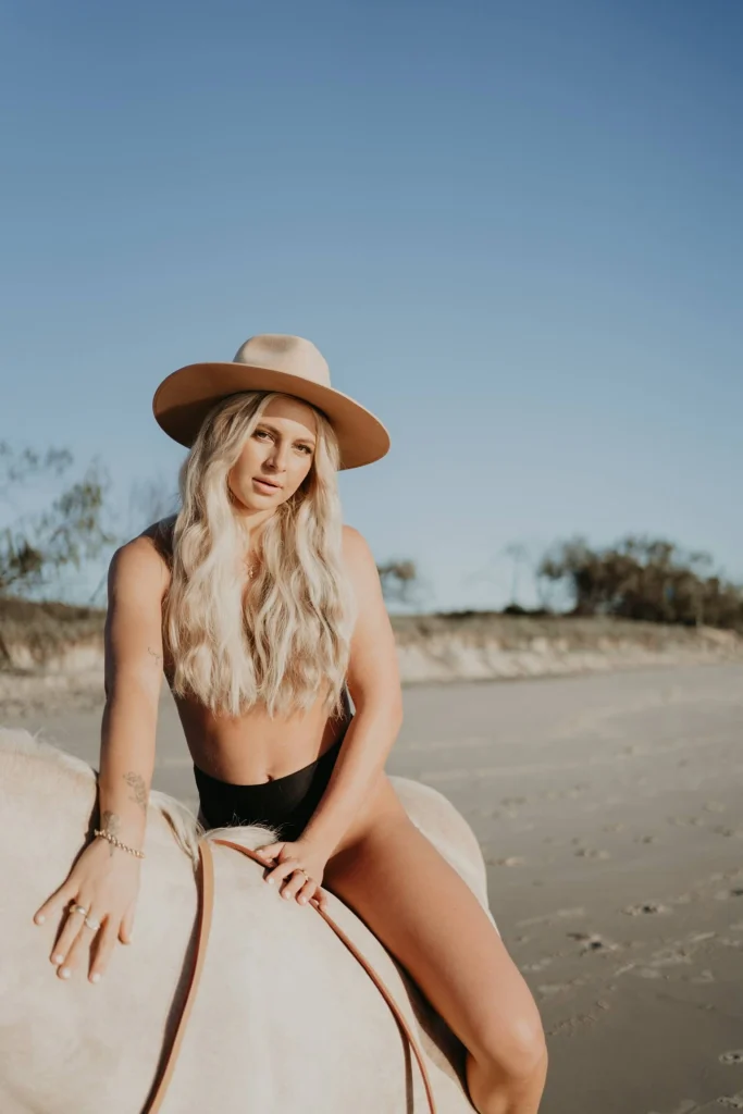 Blond woman sitting on top of a light horse on the beach.