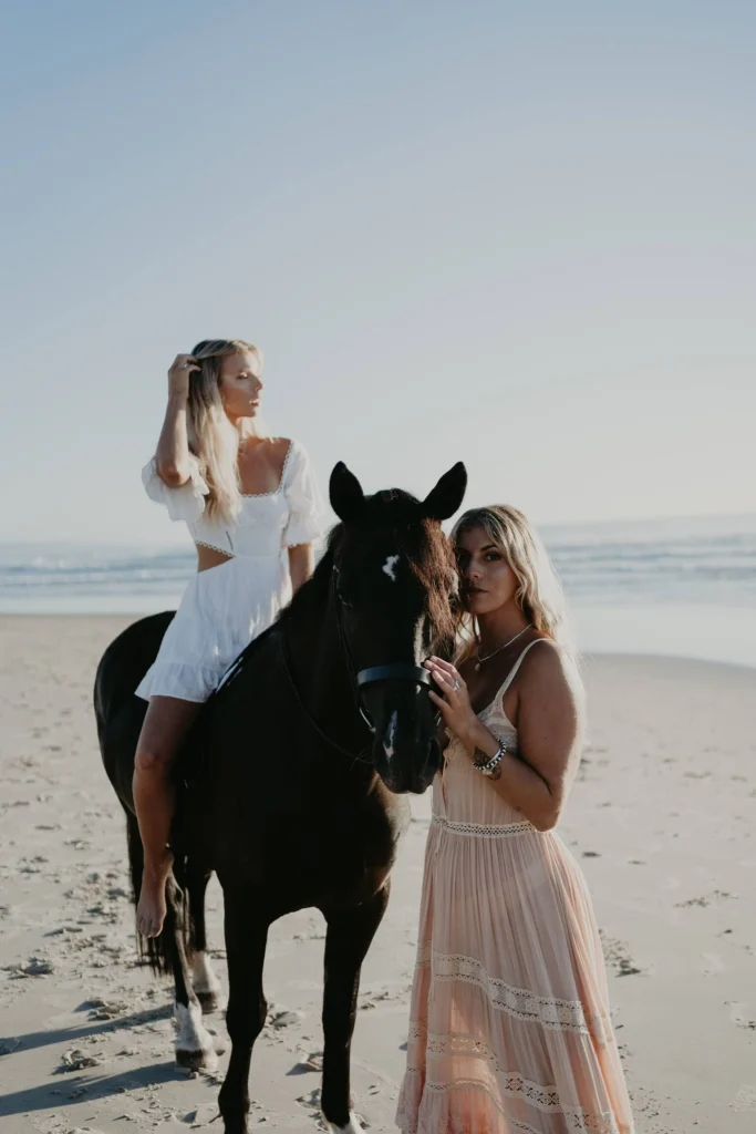 Two blond women on the beach, one sitting atop a dark horse.