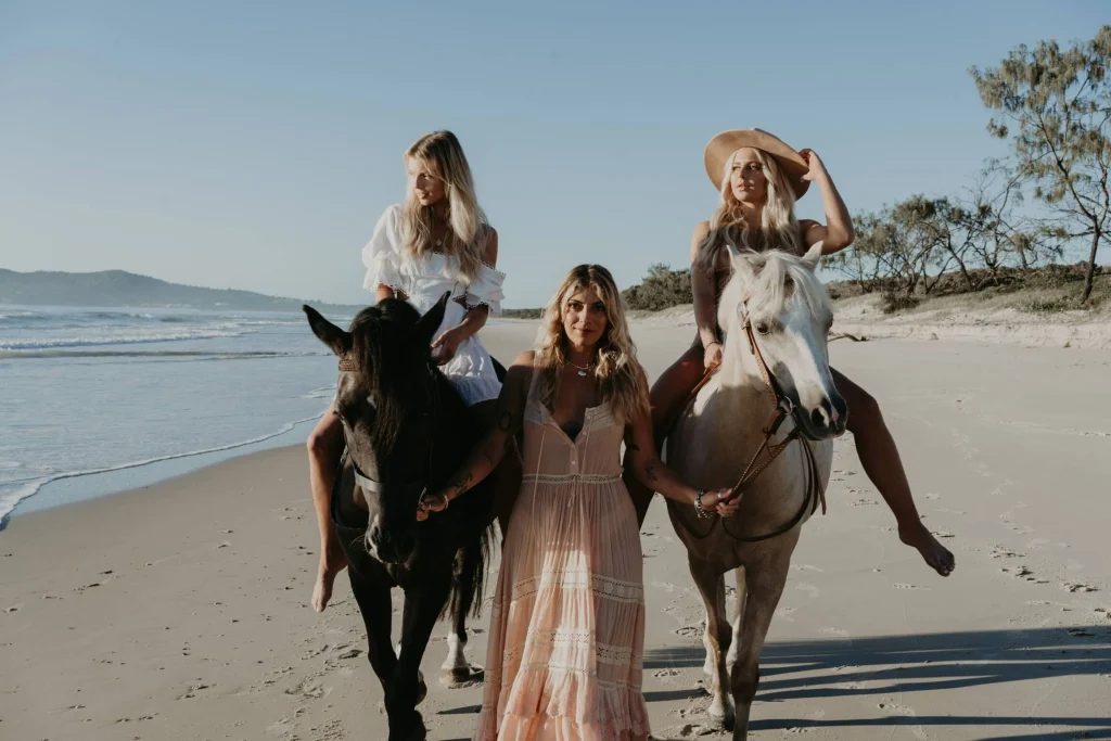 Two blond women on horses on the beach, and another walking in the centre.