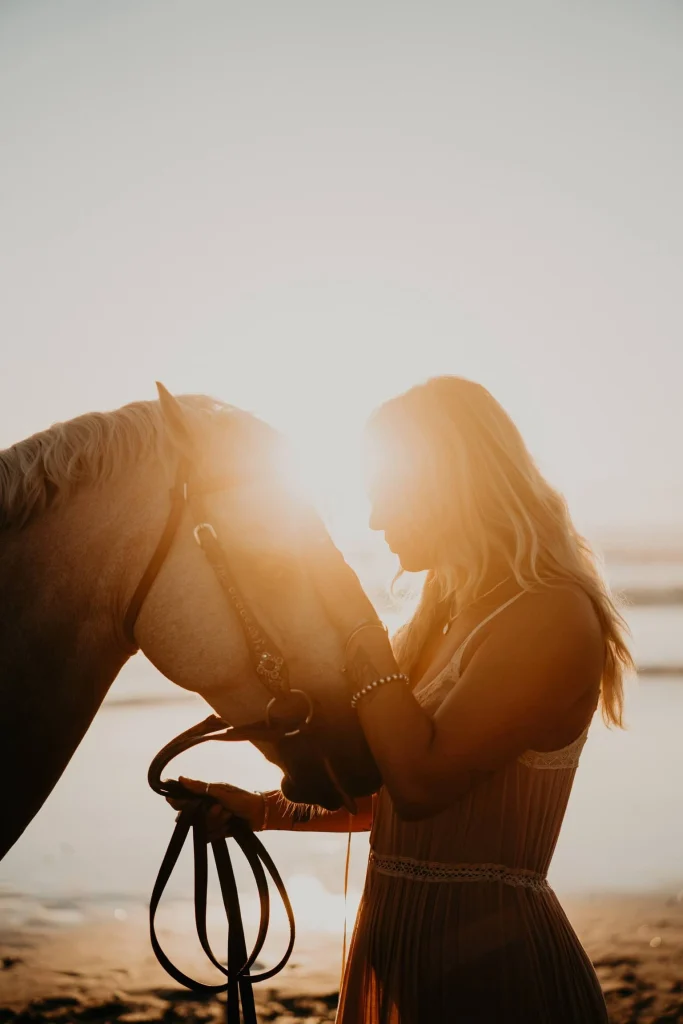 Woman standing in sunlight with her horse on the beach.