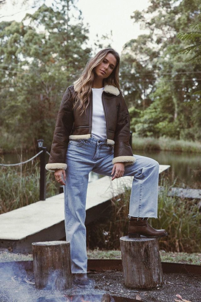 Brunette woman wearing blue jeans standing with one foot on a log around a campfire
